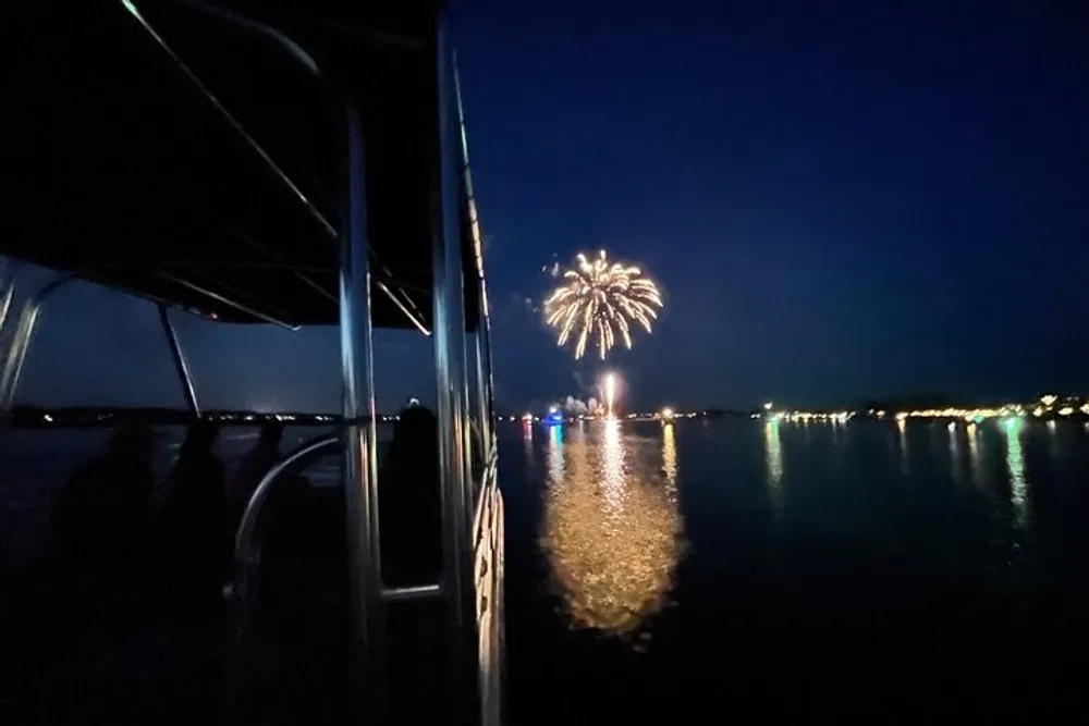 A boat is on the water at night with a view of fireworks exploding in the sky reflecting on the surface of the water
