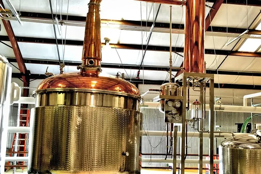 The image shows copper distillation equipment most likely used in a craft brewery or distillery, with a shiny metal exterior, gauges, and piping, set against an industrial background.
