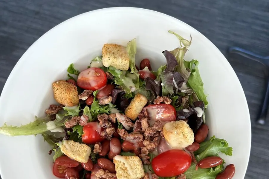 The image shows a fresh salad with mixed greens, cherry tomatoes, kidney beans, croutons, and a sprinkling of what appears to be a crunchy topping, likely nuts or bacon bits, served in a white bowl.