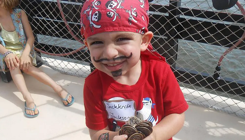 A smiling child dressed up like a pirate with a drawn-on mustache and beard holds a handful of cookies while wearing a red bandana and a red shirt