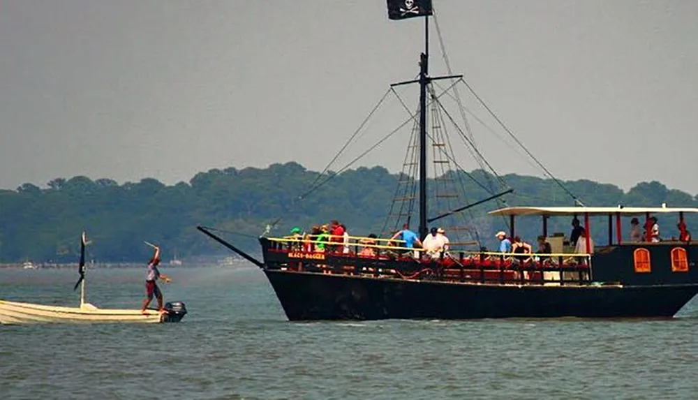 A person is standing on a small motorboat with a sail waving at a larger pirate-themed tour boat full of passengers set against a backdrop of water and trees
