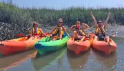 Four people in brightly colored kayaks appear to be enjoying themselves on a sunny day amidst tall grasses, projecting a sense of adventure and fun.