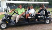 A group of people is smiling for a photo while seated in a multi-passenger golf cart labeled 