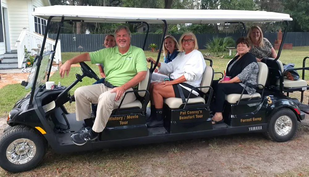 A group of people is smiling for a photo while seated in a multi-passenger golf cart labeled Pat Conroys Beaufort Tour on what looks like a guided tour
