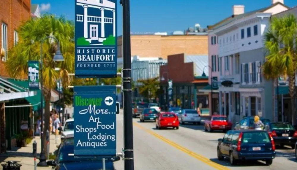 The image shows a vibrant street scene in Historic Beaufort with signs promoting local art food shops lodging and antiques set against a backdrop of palm trees and bright-colored buildings