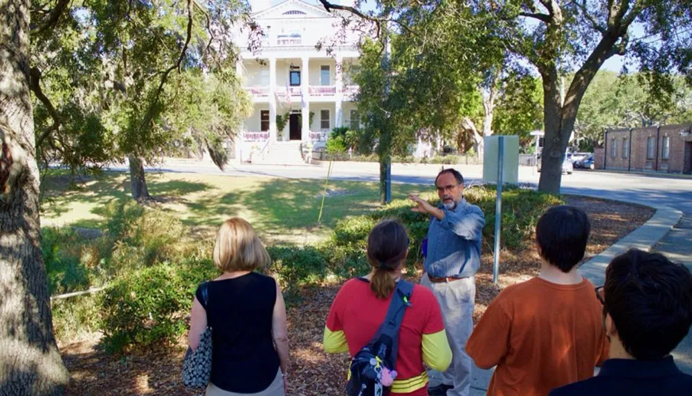 A tour guide is gesturing and explaining something to a group of attentive listeners in front of a large historic house surrounded by trees