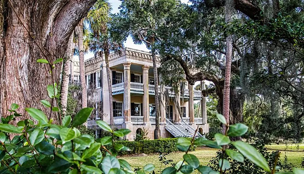 This image depicts a stately historic mansion with grand double-decker porches flanked by mature trees and lush greenery