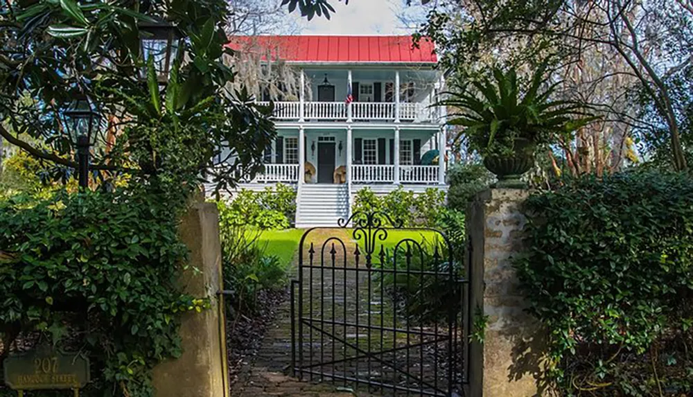 The image shows a classic two-story white house with red roofing and double porches partially obscured by lush greenery and an ornate black gate at the entrance