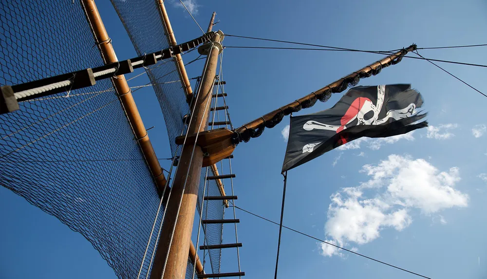 A pirate flag flutters atop a ships mast against a blue sky with some white clouds