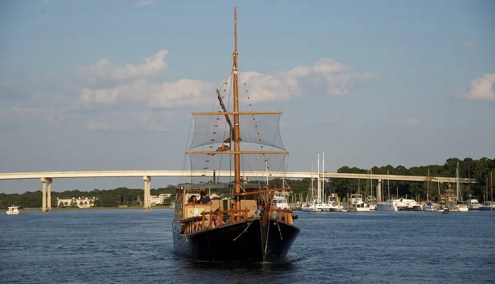 A tall ship with its sails lowered is cruising near a bridge over a body of water with other boats in the background