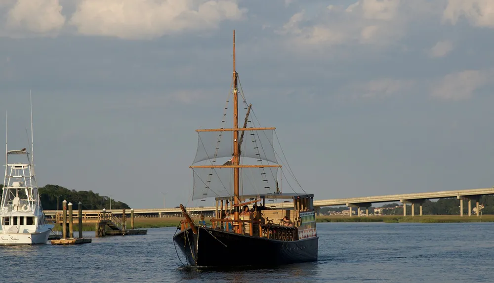 A wooden sailing ship resembling a historical or pirate vessel cruises on serene waters near a dock with a bridge in the background
