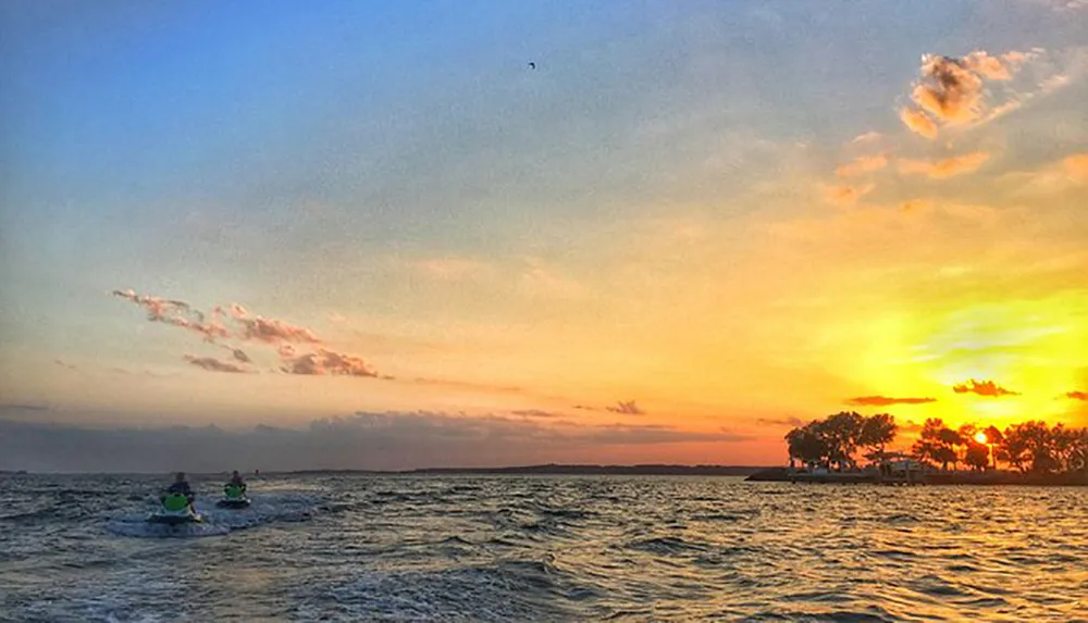 Two people are jet skiing on the water during a picturesque sunset with vibrant colors in the sky