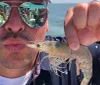A person is playfully posing with a shrimp close to their lips as if to mimic a kiss wearing sunglasses that reflect other people in a sunny outdoor setting