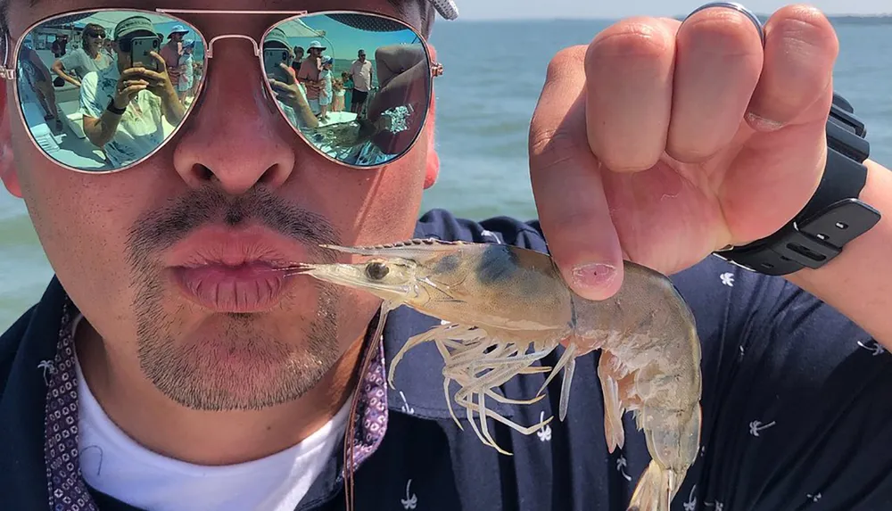 A person is playfully posing with a shrimp close to their lips as if to mimic a kiss wearing sunglasses that reflect other people in a sunny outdoor setting
