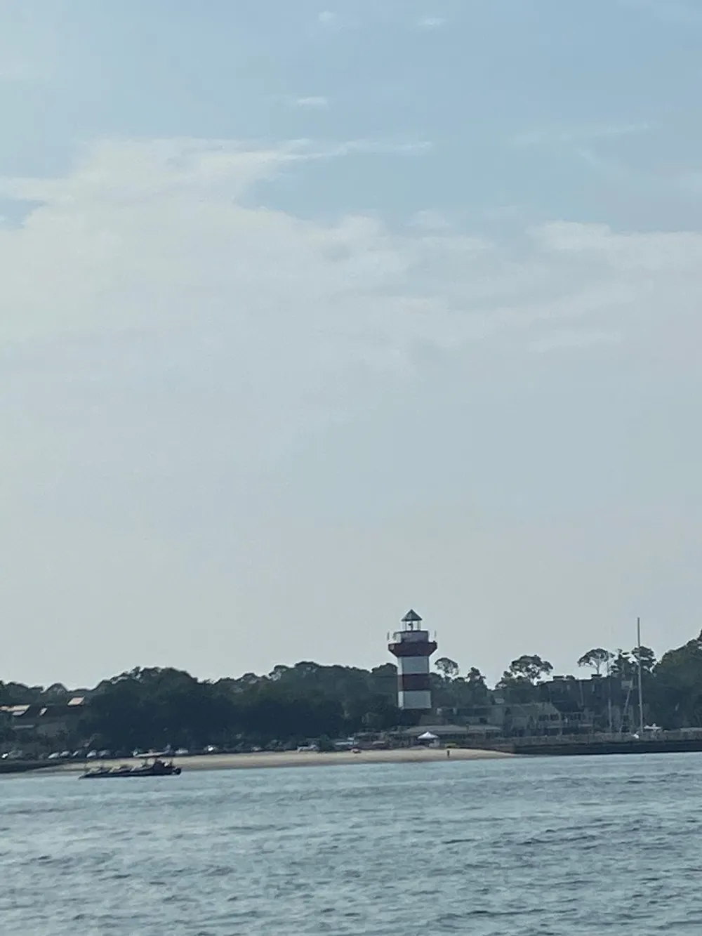The image shows a red and white lighthouse overlooking a calm body of water with a boat nearby and a backdrop of trees and a blue sky peppered with clouds