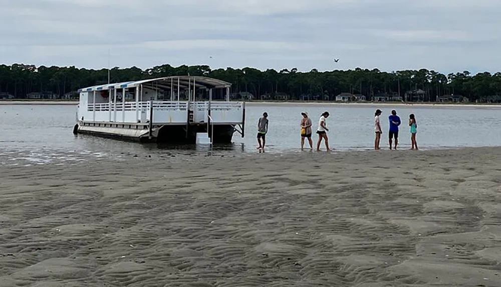 A group of people stands on a sandy shore near the waters edge with a boat docked nearby against a backdrop of trees and a cloudy sky