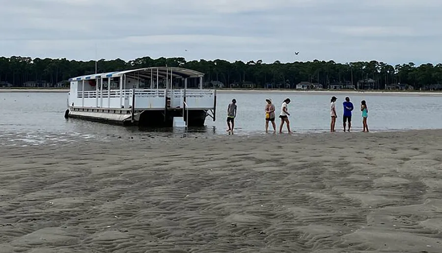 A group of people stands on a sandy shore near the water's edge, with a boat docked nearby, against a backdrop of trees and a cloudy sky.