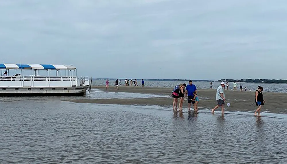 People are walking on a sandbar near a docked boat with water on either side under an overcast sky