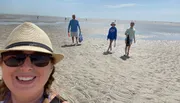 A person is taking a selfie on a sunny beach with three other people in the background walking on the sand, displaying a casual and leisurely atmosphere.