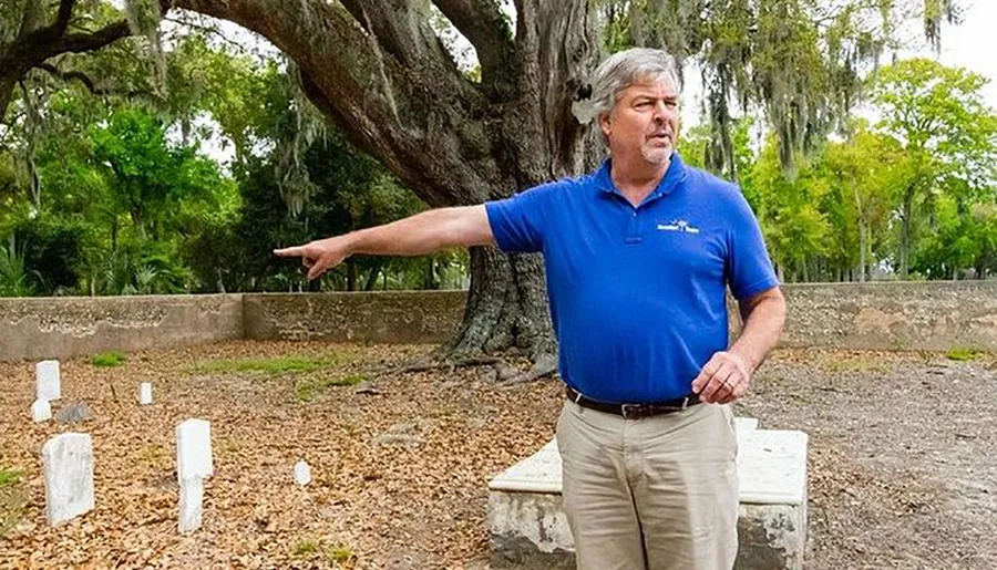 A person in a blue shirt is pointing towards the left, standing near small grave markers under a large tree draped with Spanish moss.