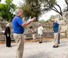 A guide wearing a blue shirt is gesturing and speaking to a small group of visitors at an outdoor historical site with stone structures and trees