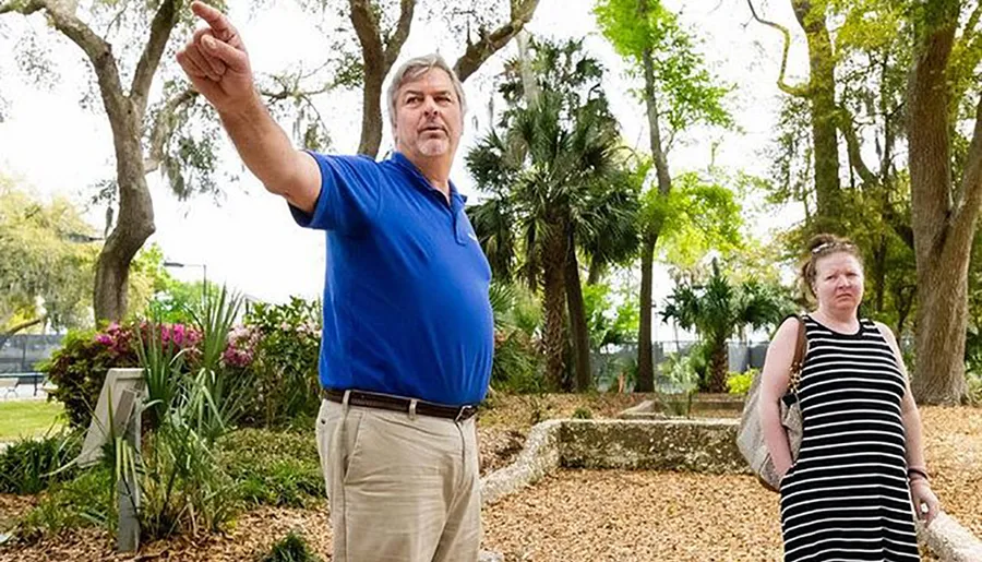 A man in a blue shirt is pointing at something while a woman in a striped dress looks on, both standing in a park with trees and flowering plants in the background.