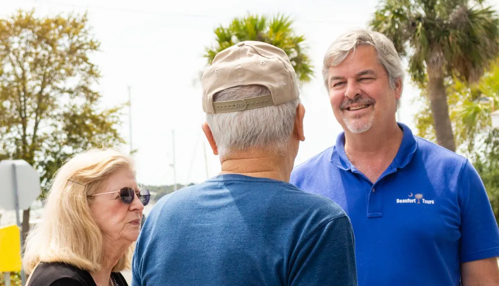 A man in a blue polo shirt with Beaufort Tours embroidered on it is smiling and conversing with an older couple outdoors