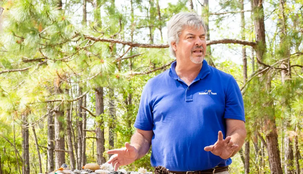 A man in a blue shirt labeled Beaufort Tours is gesturing while speaking outdoors with pine trees in the background