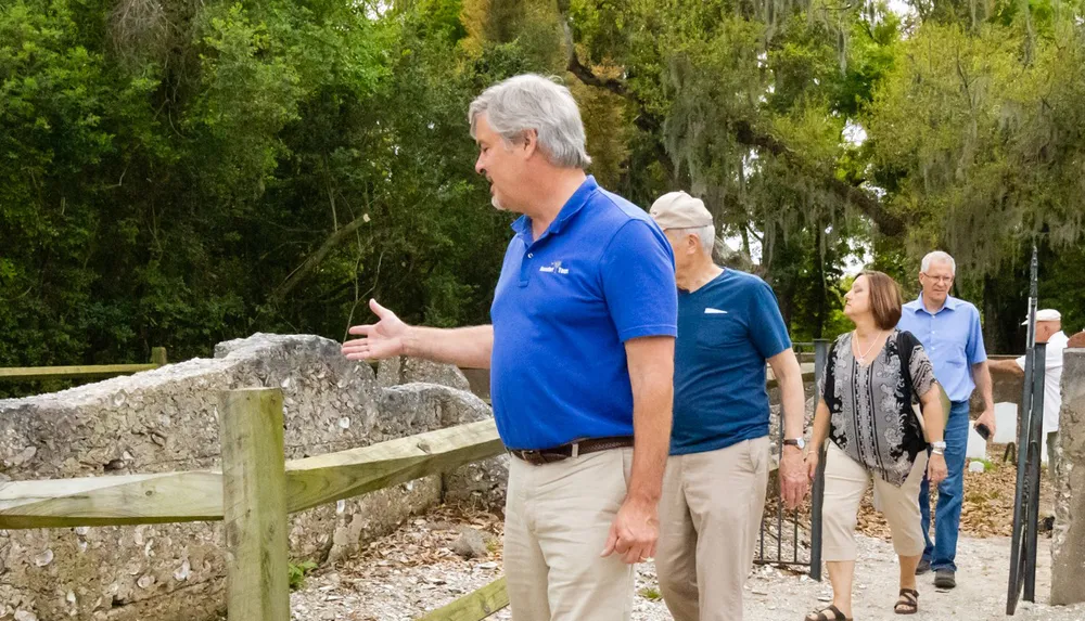 A man in a blue shirt appears to be explaining something with a gesture as others listen while walking through an outdoor area with trees and a wooden fence