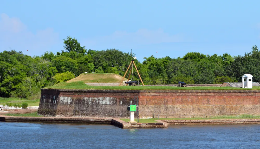 The image shows a historic fortification with grassy embankments and antique cannons overlooking a body of water under a clear blue sky