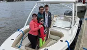 Four people are smiling for a photo on a moored white motorboat, with water and other boats visible in the background.