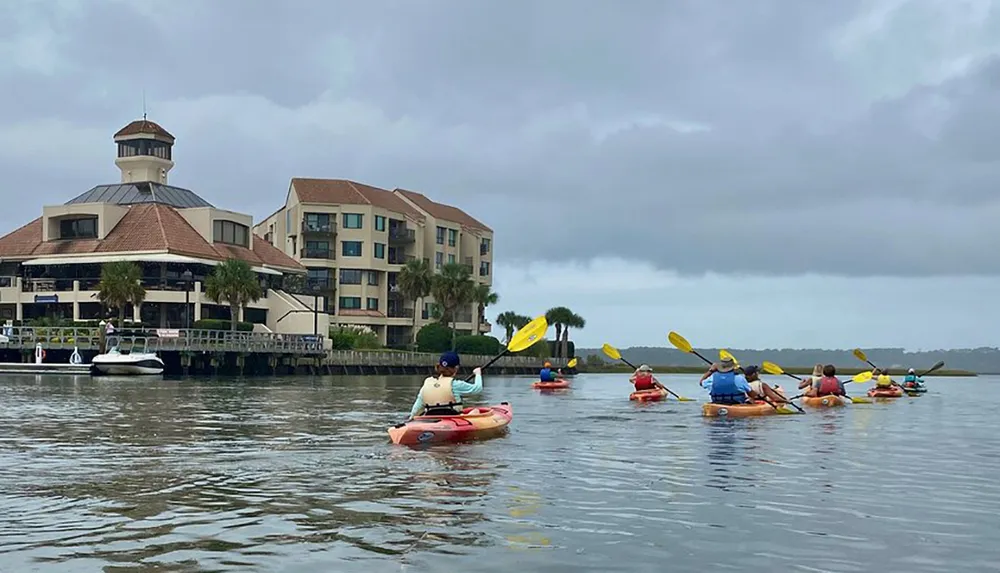 A group of people are kayaking in calm water near a coastal building complex under overcast skies