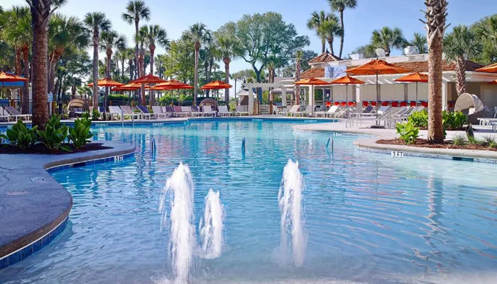 The image shows a serene resort pool area with water fountains surrounded by palm trees and loungers under orange umbrellas