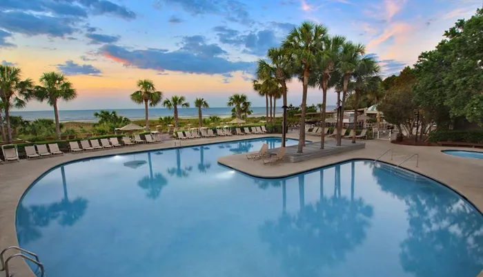 The image shows a tranquil poolside area with loungers and palm trees during twilight overlooking a serene beachscape