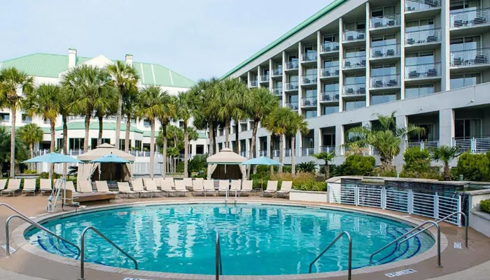 The image shows a serene hotel pool area flanked by loungers and surrounded by palm trees with a multi-story hotel building in the background