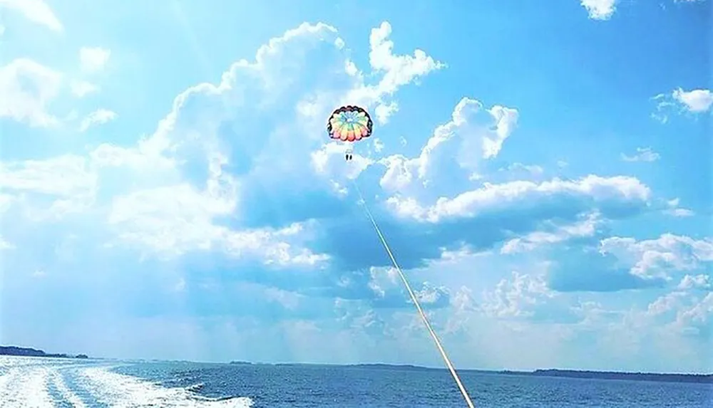 A colorful parachute is soaring in the sky with two people parasailing over a body of water on a sunny day with picturesque clouds in the background
