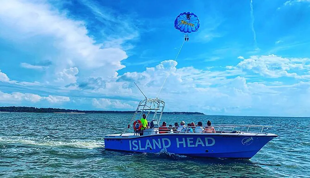 A person is parasailing high above the ocean towed by a boat filled with passengers under a partly cloudy sky