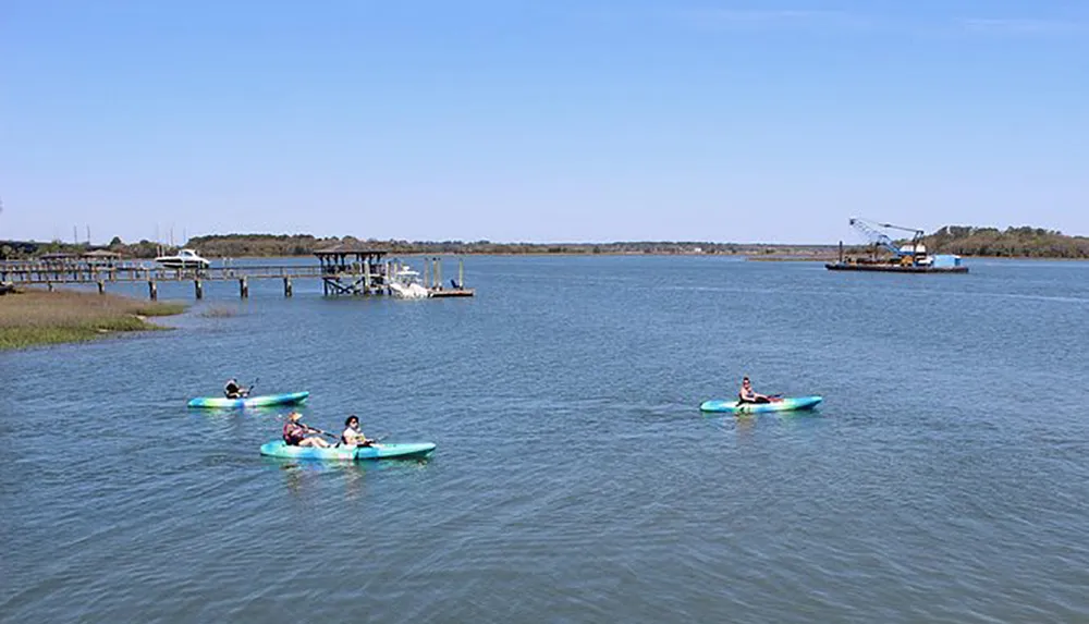 Three individuals are kayaking on a calm body of water with a pier and industrial machinery in the background