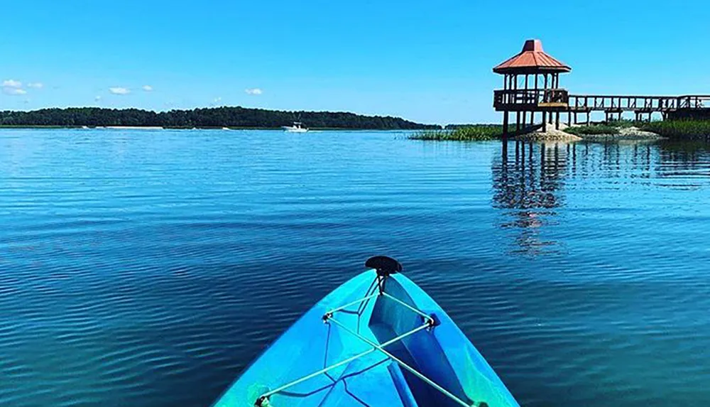 A view from a blue kayak heading towards a gazebo on a pier over calm waters with a boat and treeline visible in the distance