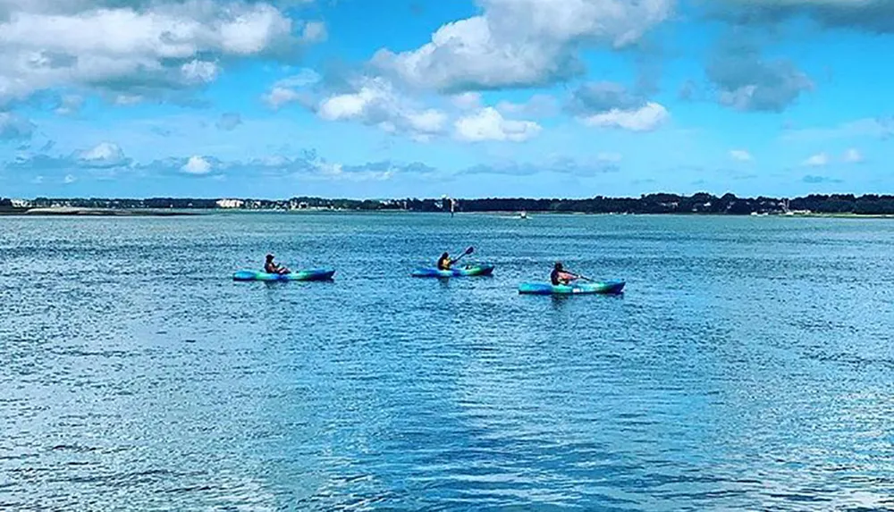 Three people are kayaking on a calm blue water body under a partly cloudy sky