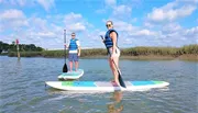 Two people are paddleboarding on calm water with grassy banks in the background.