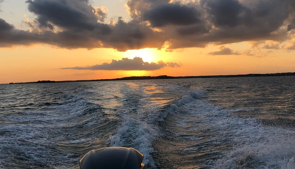 A boat creates a frothy wake on the water as the sun sets behind clouds on the horizon