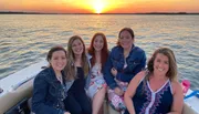 Five smiling individuals are enjoying a scenic boat ride during a beautiful sunset over a calm sea.