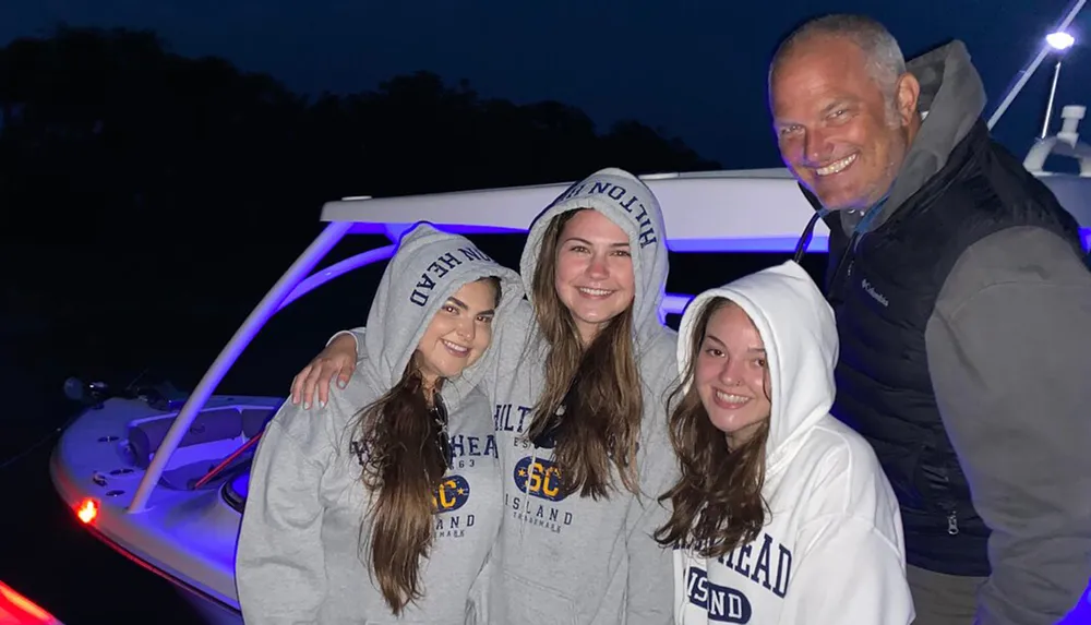 Three smiling young women wearing matching hoodies and a man are posing together on a boat at dusk with ambient lighting
