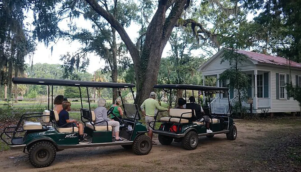 A group of people are riding in golf carts near a traditional house surrounded by greenery and Spanish moss-draped trees