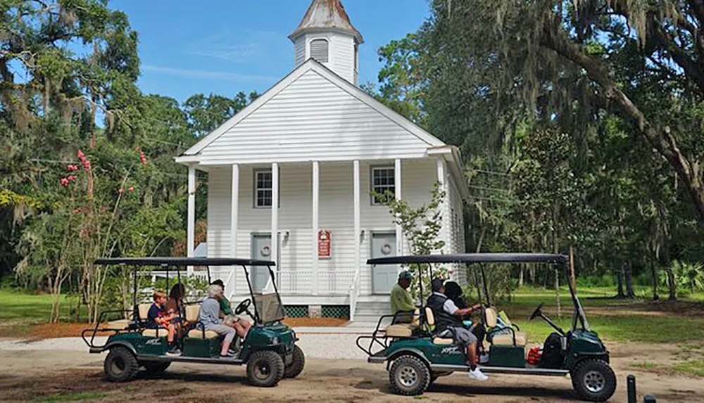 Visitors in golf carts are parked in front of a quaint white church surrounded by greenery under a blue sky
