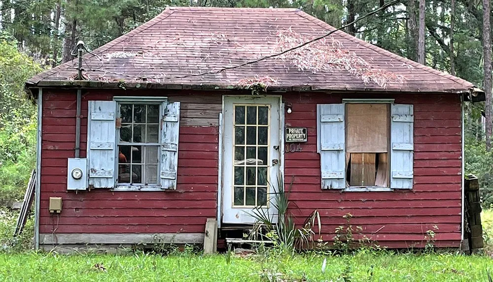 This is an image of a small dilapidated red house with boarded-up windows faded shutters and a Private Property sign on the front door set against a background of overgrown vegetation and trees