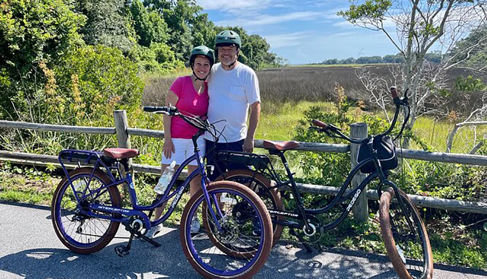 A smiling couple in helmets stands beside two bicycles on a sunny day with greenery in the background