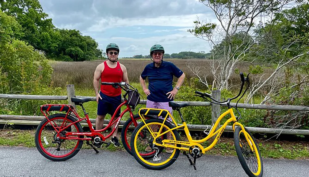 Two individuals in cycling gear are standing beside their red and yellow bicycles with a natural backdrop possibly taking a break from a bike ride