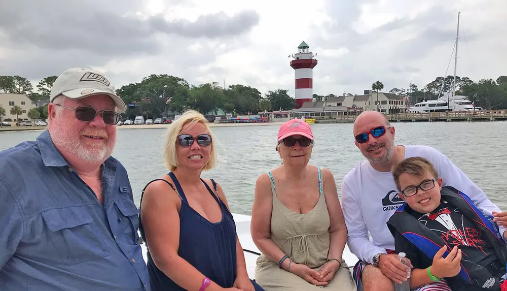 A group of five people are smiling and posing for a photo on a boat with a picturesque lighthouse and waterfront buildings in the background